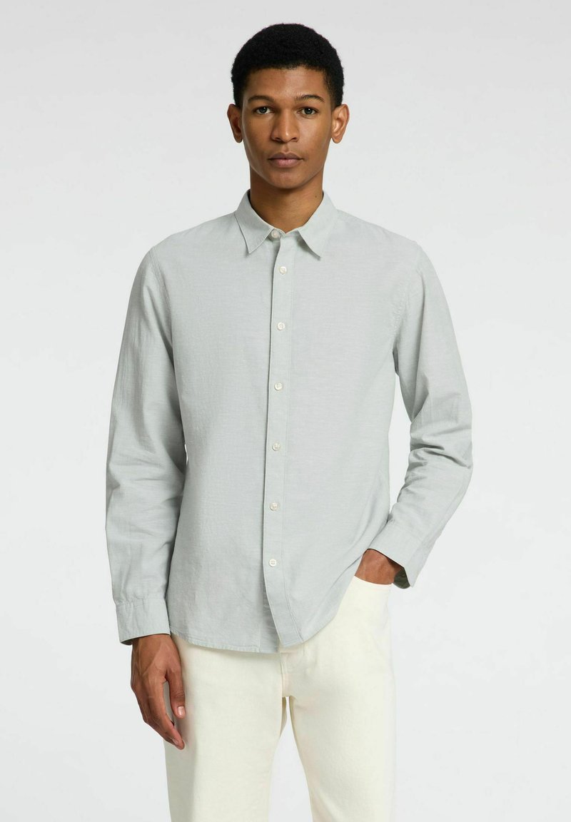 Lichtgrijze langemouwen shirt met een knoopsluiting aan de voorkant, puntige kraag en een gladde textuur. Model draagt crèmekleurige broek.