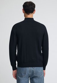 Svart stickad tröja med h ög hals, långa ärmar och ribbat avslut. Bärs över ljusblå jeans. Enkel design, mjuk textur.