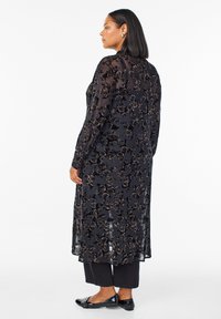 Črna dolga obleka z dolgimi rokavi s prosojnim prekrivalom, ki ima cvetlični vzorec v temnih tonih, v paru s črnimi hlačami in ravnimi čevlji.