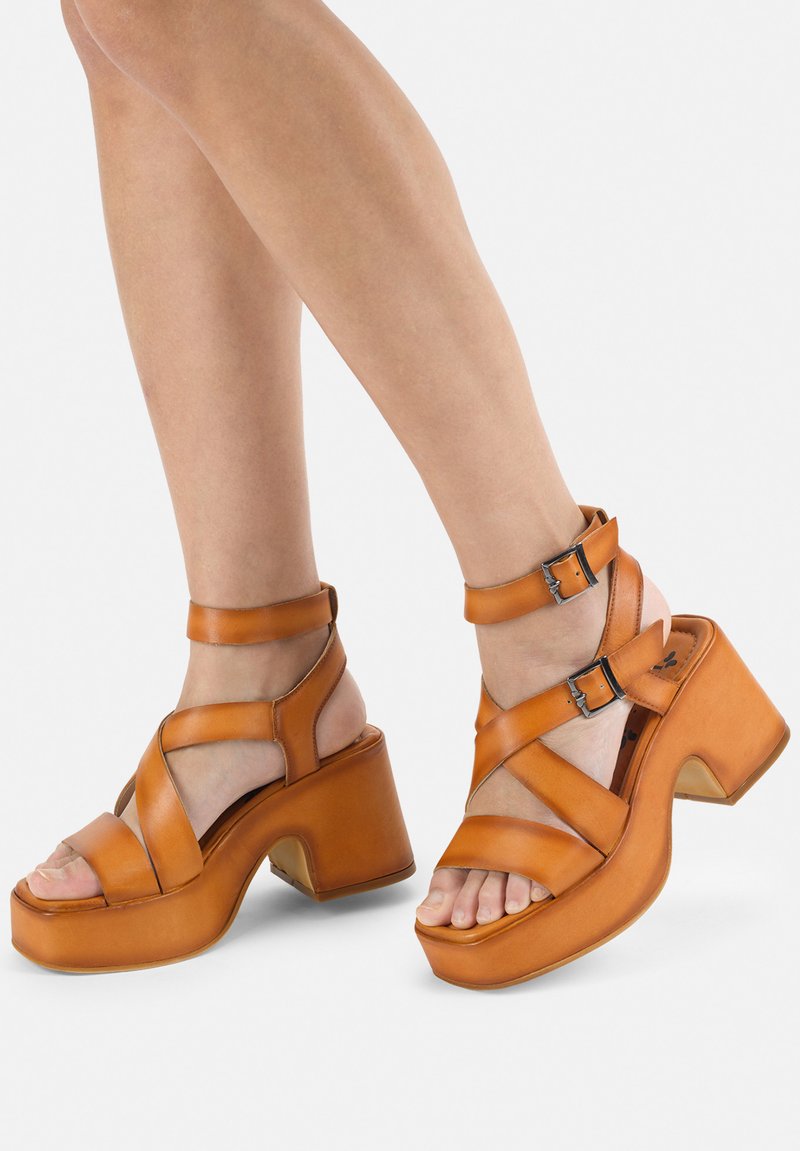 Brown leather platform sandals with crisscross straps, buckle accents, and an angular heel. Open-toe design showcasing toes.