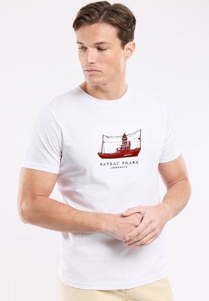 Jeune homme aux cheveux courts bruns portant un t-shirt blanc avec un graphisme de navire rouge et le texte « Bateau Phare ArmorLux ».