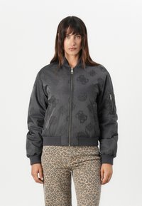 Chaqueta bomber negra con patrones tonales, cierre de cremallera, puños de canalé y un bolsillo en la manga izquierda. Combinada con pantalones de estampado de leopardo.