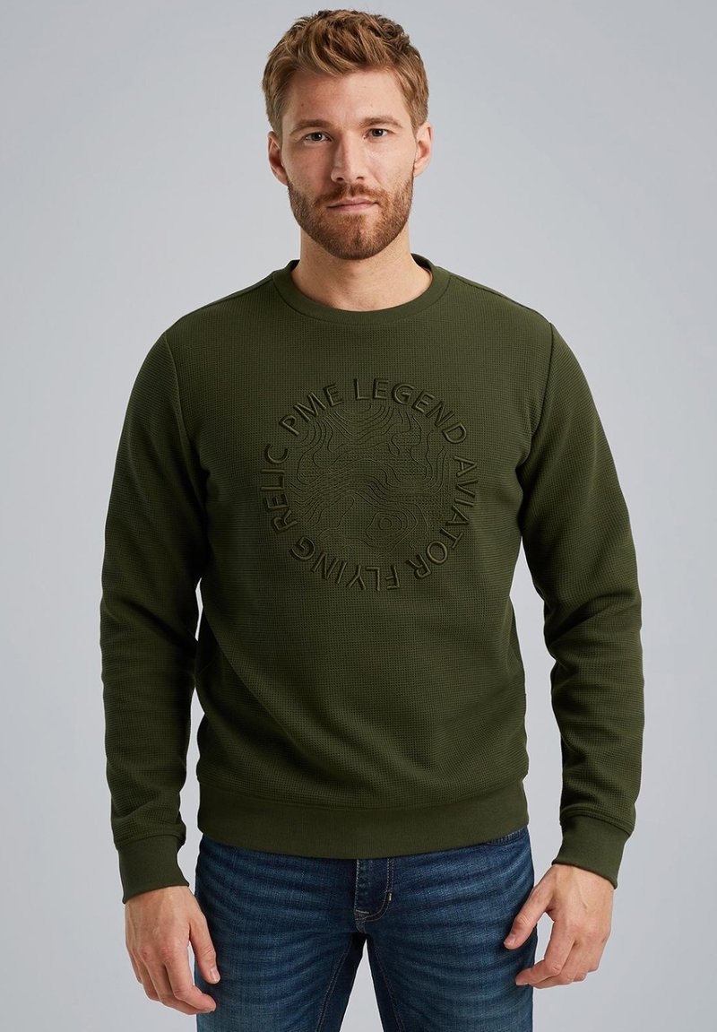 Sweatshirt texturé vert olive avec des poignets côtelés, col rond et détail de logo embossé sur la poitrine. Associé à un jean bleu.