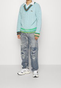 Blå kabelstickad tröja med gröna, orangea och mönstrade accenter, kombinerad med slitna ljusblå jeans och vita sneakers.