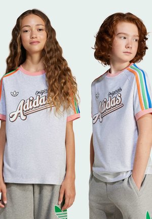 Twee jongeren dragen grijze Adidas Originals shirts met roze bies en regenboogstrepen op de mouwen, gecombineerd met grijze broeken tegen een effen achtergrond.