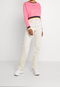 Růžová krátká mikina s modrými a oranžovými pruhy na manžetách, doplněná krémovými džíny s rovnými nohavicemi a bílými teniskami.