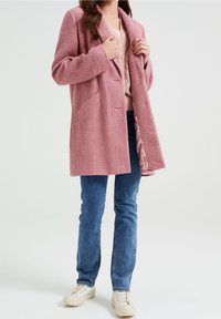 Rosa texturerad jacka med slagkrage, två framfickor och öppen front, bärs över en beige topp och blå jeans.