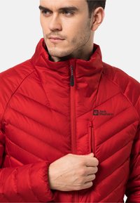 Rote gepolsterte Jacke mit hohem Kragen, ausgestattet mit einer Reißverschlusstasche und diagonalen Steppmustern. Logo auf der oberen linken Brustseite angebracht.