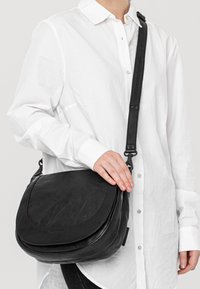 Borsa a tracolla in pelle nera con flap curvo, tracolla regolabile e texture liscia. Indossata su una camicia bianca per un contrasto.