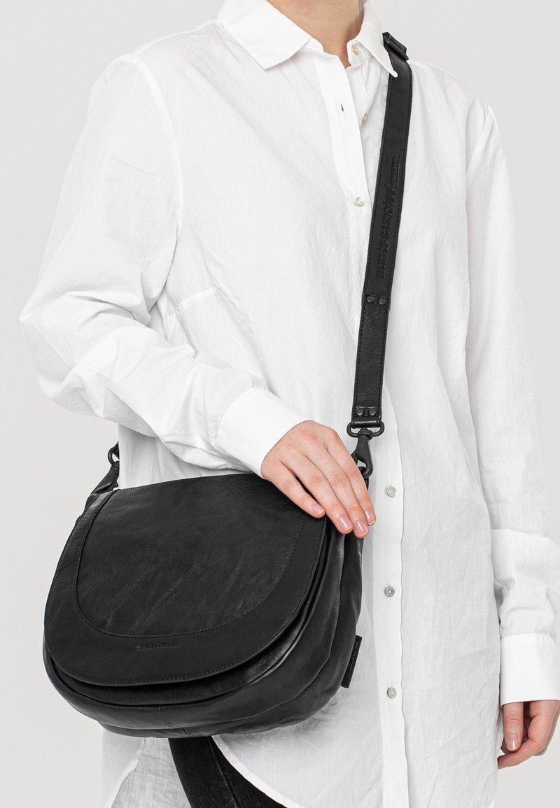 Borsa a tracolla in pelle nera con flap curvo, tracolla regolabile e texture liscia. Indossata su una camicia bianca per un contrasto.