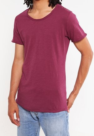 Osoba ubrana w jednolity bordowy t-shirt z krótkim rękawem i jasnoniebieskie dżinsy, stojąca na białym tle.