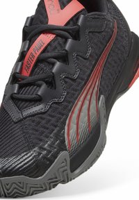 Zapato deportivo negro con materiales de malla y sintéticos, que presenta acentos rojos, superposiciones texturizadas y una suela con un patrón para tracción.