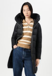 Zwarte pufferjas met een capuchon en een ritssluiting. Draag over een tan gestreepte gebreide trui. Model heeft de handen in de zakken, gecombineerd met blauwe jeans.