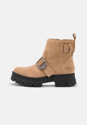 Botín de ante color canela con suela de goma negra, detalle de hebilla en el costado y logo de UGG. Presenta una lengüeta en la parte posterior para facilitar el calzado.