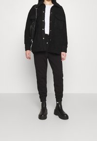 Svart texturerad ytterjacka med knapp fram och bröstfickor, ihop med svarta joggers och kraftiga svarta ankelskor.