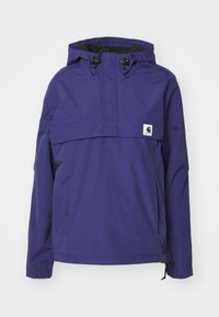 Chaqueta cortavientos morada con capucha, bolsillo frontal, cordones ajustables, puños elásticos y un pequeño parche con el logo de Carhartt en el pecho.