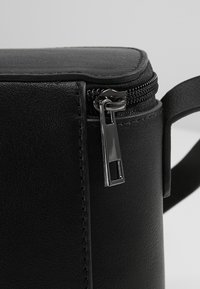 Sac en cuir synthétique noir de forme arrondie, avec une fermeture éclair métallique et une surface plate et lisse. Comprend une sangle latérale pour le porter.