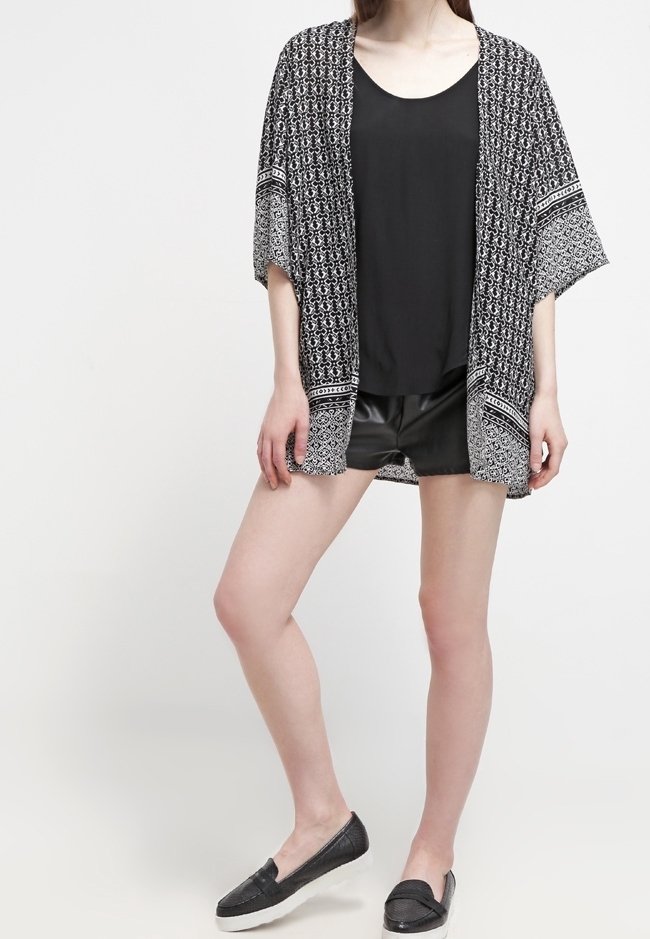 Kvinna som står klädd i svart mönstrad kimono, svart ärmlös topp, svarta lädershorts och svarta slip-on skor mot vit bakgrund.