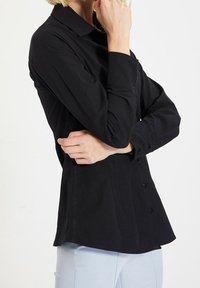 Persona che indossa una camicia nera con bottoni e maniche lunghe, colletto, braccia incrociate e pantaloni chiari su uno sfondo semplice.