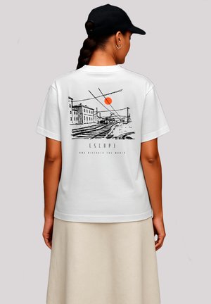 Woman wearing white t-shirt with black and orange railway track sketch and "ESCAP" text, paired with a beige skirt and black baseball cap.