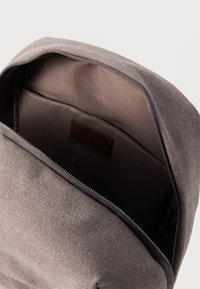 Εσωτερικό γκρίζας υφασμάτινης τσάντας πλάτης με λείο επένδυση. Διαθέτει δερμάτινη ετικέτα και κλείσιμο με φερμουάρ. Ορατή τσέπη και δομημένος σχεδιασμός.