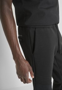 Zwarte sportbroek met een trekkoord in de taille, zijzakken en textuuraccenten op de onderbenen. Het materiaal lijkt zacht en rekbaar.