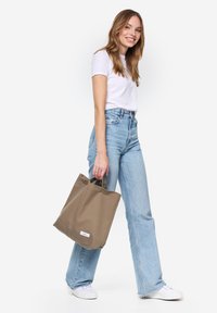 Beige vászontáska két felső fogantyúval, téglalap alakú, lapos aljú, és egy kicsi fehér címkével az oldalon. A modellen világoskék farmer és fehér póló van.