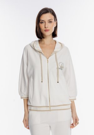 Kobieta o prostych brązowych włosach, ubrana w białą bluzę na suwak ze złotymi akcentami i dopasowane białe spodnie, stojąca na prostym białym tle.