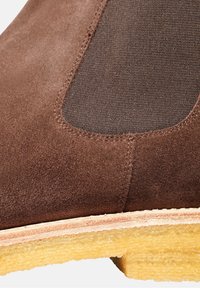 Hnědé semišové kotníkové boty s elastickou částí, texturovaným svrškem, zaoblenou špičkou a žlutou podrážkou s mírným zakřivením.