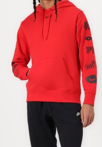 Röd hoodie med svarta grafiska mönster på ärmarna, kenguruficka, dragsko på huva och Nike-logotyp över bröstet och på ärmen.