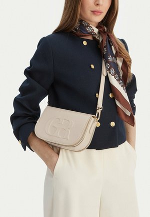 Mujer con chaqueta cruzada azul marino con botones dorados, bufanda de seda estampada, pantalones crema de talle alto y bolso bandolera beige con logo en relieve.