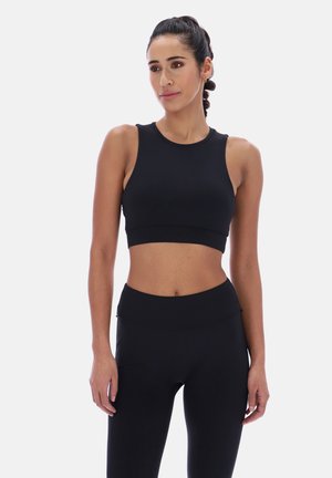 Kvinde med flettet hår iført sort, ærmeløs crop top og højtaljede sorte leggings, stående mod en ensfarvet hvid baggrund.
