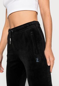 Svarta velourjoggingbyxor med sidofickor med dragkedja, elastisk midja och blåa rhinestone-logodetaljer på vänster lår.