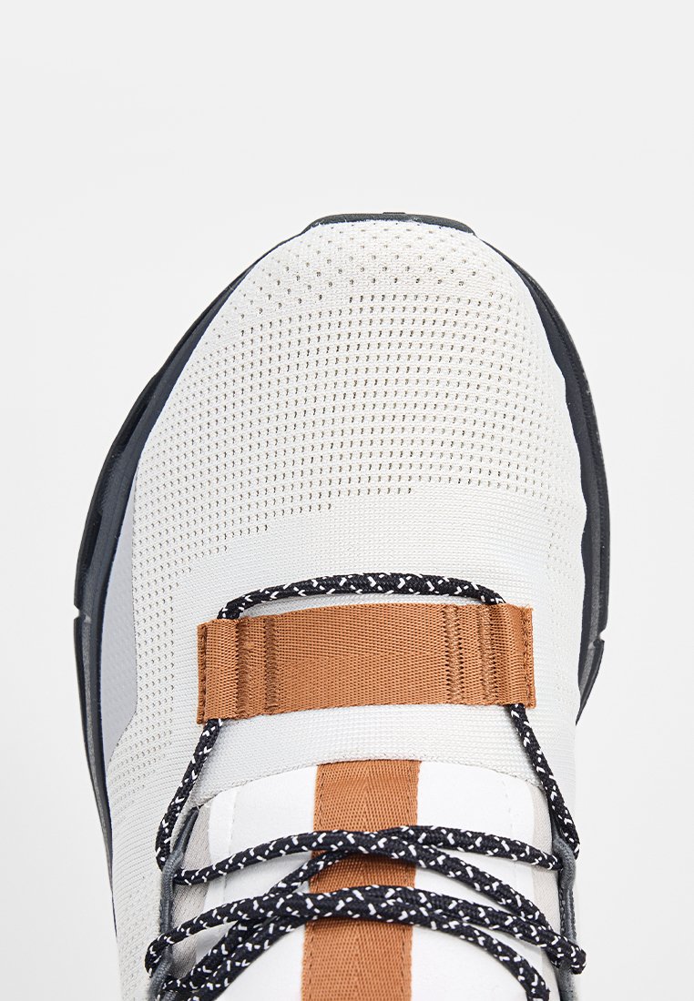 Weiße Sneaker mit atmungsaktivem Mesh-Obermaterial, schwarz-weißen gemusterten Schnürsenkeln, einem braunen Stoffriemen über der Oberseite und schwarzen Sohlenkanten.