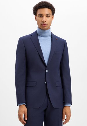 Junger Mann, der einen marineblauen Anzug und einen hellblauen Rollkragenpullover trägt, vor einem schlichten weißen Hintergrund stehend.