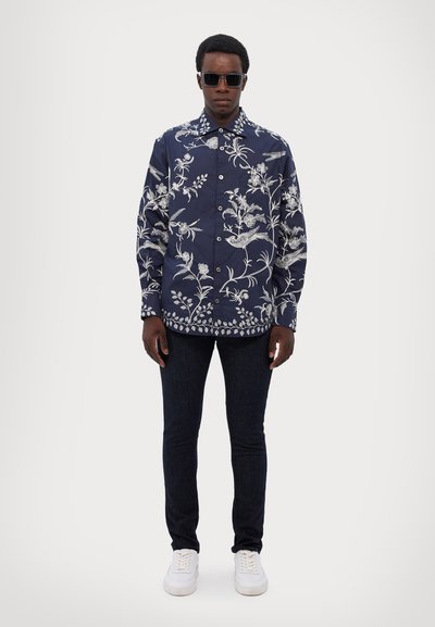 Camisa de botones azul marino con bordado floral blanco, combinada con jeans de mezclilla oscura y zapatillas blancas. Lleva gafas de sol y está en una pose de pie.