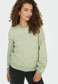 Sweat-shirt vert clair avec des ornements en forme de cœur dorés, des poignets côtelés et un col rond, associé à un jean foncé.