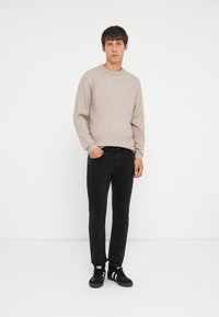 Suéter de punto beige claro con cuello redondo, combinado con jeans negros y zapatillas negras. La tela parece suave y texturizada.