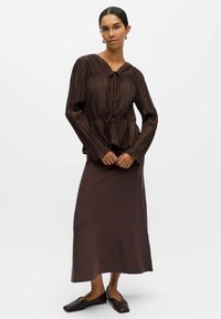 Brun plisserad blus med dragsko i midjan, kombinerad med en enfärgad brun maxi kjol. Outfiten har en mjuk textur och avslappnad silhuett.