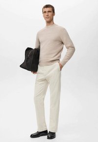 Jersey de punto beige claro, pantalones color crema, zapatos de cuero negro, sosteniendo un bolso de ante negro, de pie contra un fondo neutro.