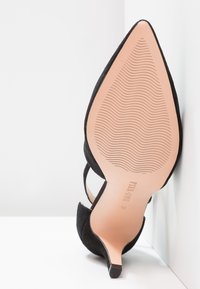 Scarpa nera in suede con tacco alto, punta affusolata e sottile tacco a stiletto. La suola è di un beige chiaro con un motivo testurizzato.