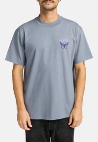 Camiseta de algodón azul claro con cuello redondo, que presenta un gráfico de mariposa púrpura y el texto "RVCA" en el lado izquierdo del pecho. Mangas cortas.