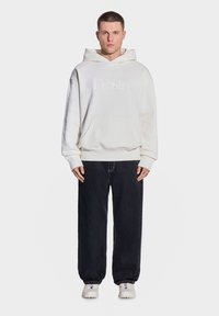 Biała bluza z kapturem z haftowanym logo, dużą kieszenią z przodu i luźnym krojem, w zestawie z ciemnymi dżinsami i jasnymi sneakersami.