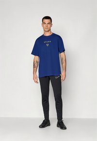 Blauw T-shirt met de tekst "SPURS" en logo, gecombineerd met zwarte slim-fit broek met een klein Nike-logo, gedragen met zwarte sneakers.