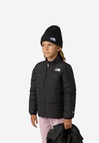 Černá prošívaná bunda s předním zipem, vysokým límcem a logovými detaily; na hlavě má černou pletenou čepici. Světlé růžové kalhoty a černou bundu drží v ruce.