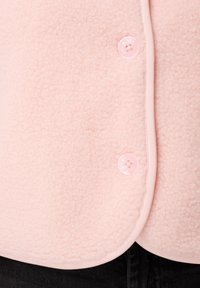 Lichtroze fleecejack met een gladde textuur, voorzien van een ronde kraag en roze knoopsluitingen. Afgewerkt met een bijpassende bies.