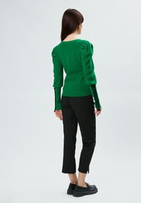 Grüner Strickpullover mit Ballonärmeln, kombiniert mit schmalen schwarzen Hosen. Der Pullover hat schwarze Akzentdetails an den Ärmeln. Schwarze Plateauschuhe.