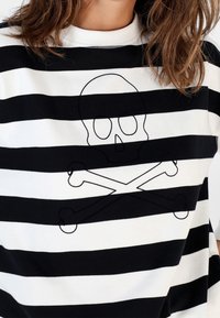 Femme portant un t-shirt à rayures horizontales noires et blanches avec un design de crâne et os croisés brodé sur la poitrine.