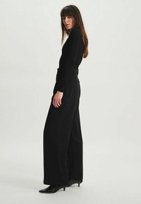 Haut noir à manches longues associé à un pantalon noir à jambes larges, avec une taille haute et une silhouette élégante. Les chaussures sont des escarpins noirs pointus.