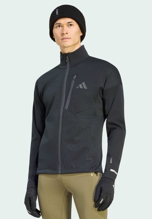 Uomo che indossa un berretto nero, giacca nera con zip e tasca sul petto, guanti neri e pantaloni beige, in piedi su sfondo chiaro.